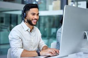 快乐的年轻男性销售代理通过耳机与客户交谈，并在计算机工作站工作